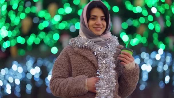 Leende vacker kvinna tittar på kameran vänder sig till julbelysning i bakgrunden. Glad säker Mellanösterndam stående i urban stad njuter av nyårsfirande poserar. — Stockvideo