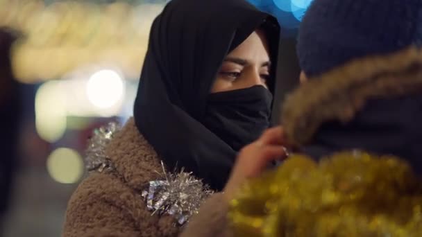 Primer plano hermosa madre musulmana abrazando niño en la noche de Navidad en la ciudad. Delgado confiado en la mujer de Oriente Medio en pañuelo para la cabeza abrazando hijo en Año Nuevo al aire libre. Amor y felicidad familiar. — Vídeos de Stock