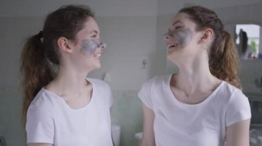 Yüzünde kozmetik maske olan birbirine gülen iki güzel kadın kameraya gülümsüyor. Sabah banyoda poz veren beyaz kız kardeşlerin portresi..