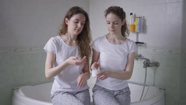 Sorelle gemelle in pigiama sedute sulla vasca da bagno che applicano crema idratante sulle mani che sorridono. Ritratto medio due giovani donne caucasiche belle fiduciose al mattino in bagno. — Video Stock