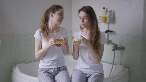 Идентичные сестры-близнецы разговаривают сидя на ванне с чаем и кофе в чашках. Улыбаясь позитивным молодым кавказским красивым женщинам в пижамах, наслаждаясь утренним разговором в ванной комнате, делясь секретами. — стоковое видео