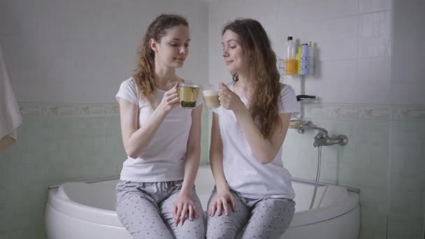 Twee zelfverzekerde tweelingzussen die 's morgens thee en koffie drinken in de badkamer. Portret van slanke aantrekkelijke blanke jonge vrouwen genieten weekend thuis met drinken. — Stockvideo