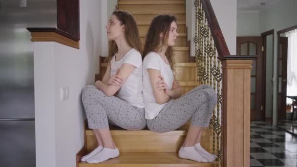 Dissatisfied argued twin sisters in pajamas sitting on stairs back to back. Side view portrait annoyed irritated Caucasian young identical women thinking. Siblings conflict. — Stock Video