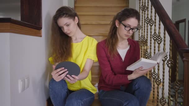 Inteligentna młoda kobieta w okularach czyta książkę siedzącą na schodach z siostrą bliźniaczką podziwiającą odbicie w lustrze. Identyczne bliźnięta kaukaskie w domu. Pojęcie różnicy indywidualnej. — Wideo stockowe