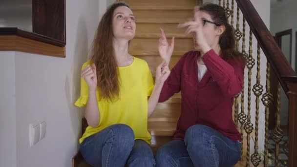Gemelos idénticos hablando de gestos explicando diferentes cosmovisiones entre sí. Retrato de mujeres jóvenes caucásicas delgadas con una individualidad diferente sentadas en las escaleras en casa charlando. — Vídeo de stock
