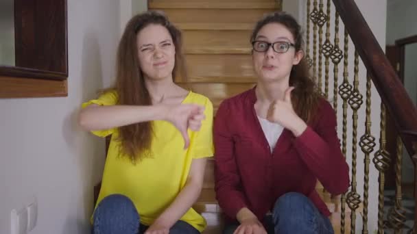 Smiling positive woman in eyeglasses gesturing thumb up as skeptic twin sister showing thumb down with sarcastic facial expression. Identical Caucasian adult twins posing sitting on stairs at home. — Stock Video