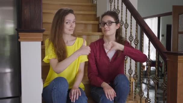 Skeptic young woman in eyeglasses showing thumb down as cheerful twin sister gesturing thumb up smiling looking at camera. Identical twins with different attitude posing sitting on stairs at home. — Stock Video