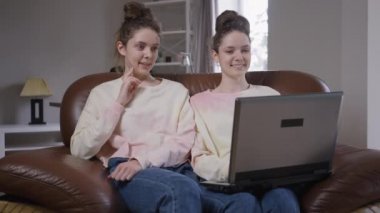 Rahat bir kanepede oturup dizüstü bilgisayarla sosyal medyada gezinen rahat ikiz kız kardeşlerin portresi. Neşeli gülümseyen güzel beyaz kadınlar evde dedikodu yapıyorlar..