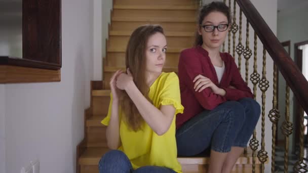Portrait of confident slim beautiful woman looking at camera touching hair with sarcastic twin sister in eyeglasses sitting at background on stairs. Narcissistic Caucasian lady with nerd sibling. — Stock Video