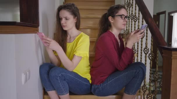 Sorelle gemelle assortite che navigano sui social media su smartphone parlando seduti sulle scale di casa al chiuso. Rilassato caucasico giovani belle donne discutendo voci. Femminilità e individualità. — Video Stock