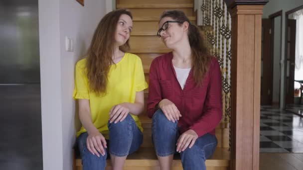 Ευτυχισμένες δίδυμες αδερφές που χαμογελούν αγκαλιασμένες κοιτάζοντας την κάμερα που κάθεται στις σκάλες στο σπίτι. Θετική όμορφη λεπτή Καυκάσια νεαρές γυναίκες θέτουν αγκαλιάζει. Η έννοια της αδελφότητας και της ευτυχίας. — Αρχείο Βίντεο