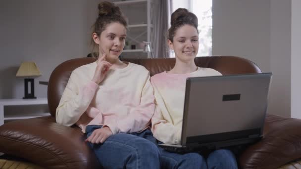 Retrato de hermanas gemelas jóvenes y relajadas sentadas en un cómodo sofá surfeando en las redes sociales en el portátil. Alegre sonriente hermosa caucásica mujeres hablando chismes en casa en interiores. — Vídeos de Stock