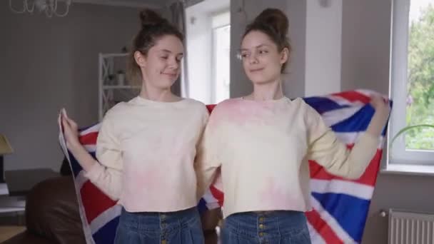 Μέτριο πλάνο χαμογελαστή δίδυμες αδελφές τυλίγοντας σε βρετανική σημαία αγκαλιάζει κοιτάζοντας την κάμερα. Πορτρέτο του ευτυχισμένη όμορφη νεαρή Καυκάσια γυναίκες ποζάρουν στο σαλόνι στο σπίτι με εθνικό σύμβολο. — Αρχείο Βίντεο