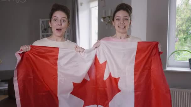 Irmãs gêmeas entusiasmadas fãs de hóquei com bandeira canadense apoiando jogo de TV na sala de estar em casa regozijando o sucesso. Visão frontal retrato alegre jovens mulheres idênticas falando sorrindo pulando em casa. — Vídeo de Stock