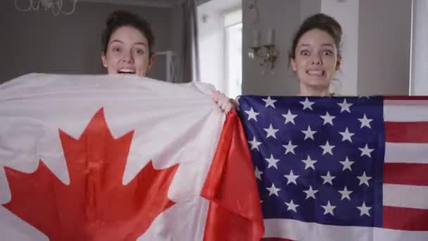 Сестры-близнецы с канадскими и американскими флагами смотрят чемпионат по телевизору дома, поддерживая хоккей или футбольные команды. Портрет двух одинаково красивых женщин, болеющих за другую страну. — стоковое видео
