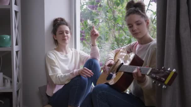 Portret van een ontspannen, mooie vrouw die zingt met een tweelingzus die thuis gitaar speelt op een vensterbank. zorgeloze blank slanke vrouwen genieten van hobby op weekend binnen. — Stockvideo