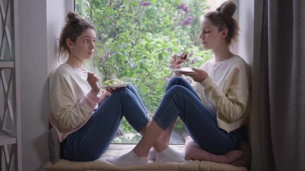 Блискучі близнюки їдять здоровий салат і нездорову солодку булочку. Широкий боковий вид Кавказькі молоді жінки з різною їжею сидять на віконному підвіконні вдома за обідом.. — стокове відео