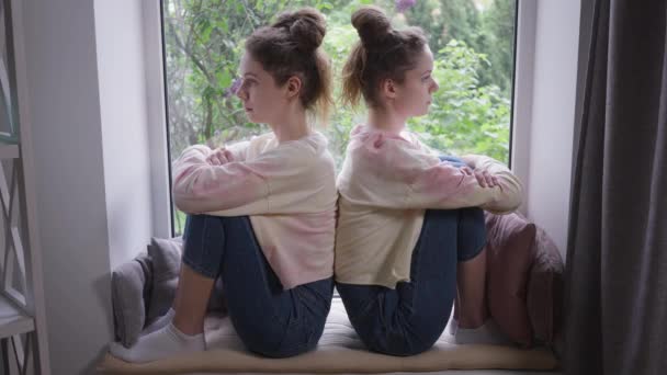 Side view to identiske unge kvinder sidder i vindueskarmen tilbage til bagtanken. Bred skud argumenterede stædige kaukasiske tvillingesøstre med krydsede hænder derhjemme indendørs. Konflikter og misforståelse. – Stock-video