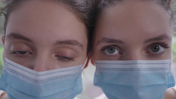 Крупным планом глаза идентичных сестер-близнецов в коронавирусных масках, смотрящих в камеру с серьезным выражением лица. Портрет стройных молодых красивых кавказских женщин, позирующих в изоляции Ковид-19. — стоковое видео