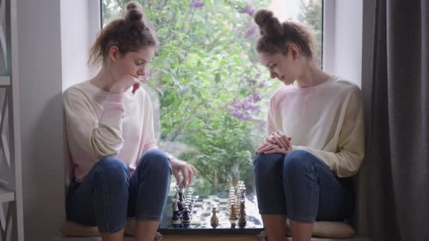 Mujer blanca hermosa inteligente jugando al ajedrez con hermana gemela sentada en el alféizar de la ventana. Hermano perdiendo barrer tablero de ajedrez dejando. Sonriente ganador confundido con expresión facial impactada. — Vídeos de Stock