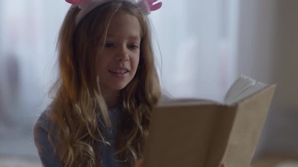 Portræt af charmerende glad lille pige læse højt sidder derhjemme indendørs. Temmelig kaukasisk barn nyder hobby om aftenen. Intelligens og livsstil koncept. – Stock-video