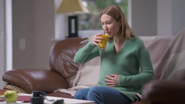 Ευτυχισμένη νεαρή έγκυος γυναίκα πίνοντας υγιεινό χυμό πορτοκαλιού χαϊδεύοντας την κοιλιά σε αργή κίνηση κάθεται σε άνετο καναπέ στο σπίτι. Πορτρέτο του χαμογελαστού Καυκάσιου αποχρεμπτικό. — Αρχείο Βίντεο