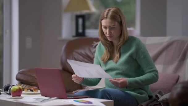 Gericht drukke zwangere vrouw die thuis papierwerk analyseert zittend met laptop op de bank in de woonkamer. Portret van geconcentreerde Kaukasische verwachting freelancer planning business idee. — Stockvideo