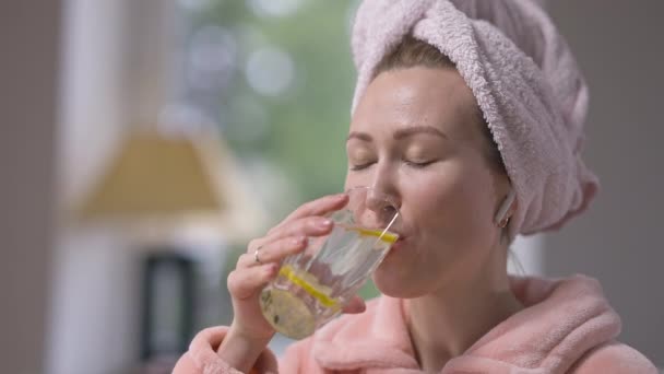 Тонкая молодая женщина пьет лимонную детоксикационную воду в замедленной съемке, наслаждаясь вкусом и глядя на улыбающуюся камеру. Счастливая позитивная кавказская дама, заботящаяся о здоровье и красоте по утрам. — стоковое видео
