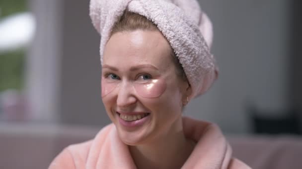 Front view laughing woman with eye patches looking at camera gesturing air kiss in slow motion. Portrait of carefree positive Caucasian lady in hair towel and bathrobe posing in the morning at home. — Stock Video