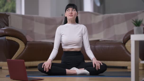 Weitschuss konzentrierte schöne schlanke Frau, die in Lotus-Pose sitzt und die Augen schließt, während sie zu Hause meditiert. Porträt von vorne selbstbewusst attraktive kaukasische Yogi im Wohnzimmer. Zeitlupe. — Stockvideo
