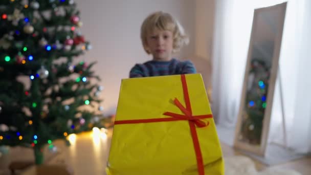 Κίτρινο τυλιγμένο χριστουγεννιάτικο δώρο με κόκκινη κορδέλα και θολό χαρούμενο αγοράκι που τεντώνει το παρόν χαμογελώντας στο παρασκήνιο. Ξέγνοιαστη χαρούμενη Καυκάσιος παιδί ποζάρουν με κουτί στο σπίτι την παραμονή της Πρωτοχρονιάς. — Αρχείο Βίντεο