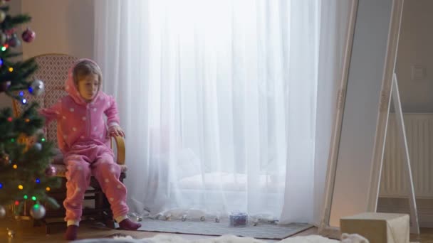 Кавказька дівчинка, що сиділа в кріслі-гойдалці, підходила до вікна і чекала на Санту ввечері вдома. Широко застрелив щасливого кавказького хлопчика в рожевому костюмі в новому році в зручному будинку.. — стокове відео