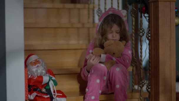 Portret smutnej białej dziewczynki siedzącej na schodach i przytulającej misia w Wigilię. Nieszczęśliwy zdenerwowany dzieciak w domu z zabawką w Nowy Rok. Koncepcja smutku. — Wideo stockowe