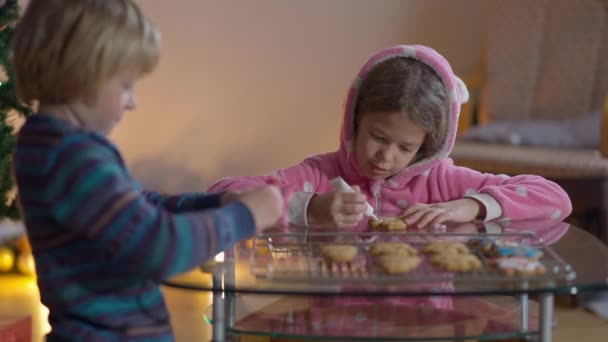 Konzentriertes Mädchen beim Dekorieren von Weihnachtsplätzchen mit Jungen drinnen. Porträt der süßen kaukasischen Schwester und des Bruders, die zu Hause an Silvester ein süßes Dessert zubereiten. Traditionskonzept. — Stockvideo