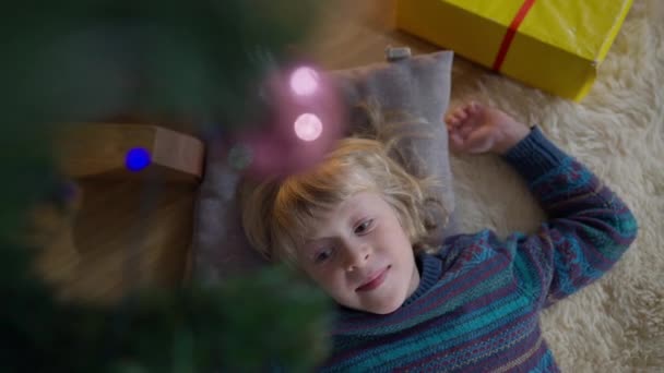 Vista superior feliz niño relajado acostado en el árbol de Navidad con el regalo de Año Nuevo sonriendo. Retrato de un niño caucásico despreocupado disfrutando de unas vacaciones en la sala de estar en casa dentro. — Vídeo de stock