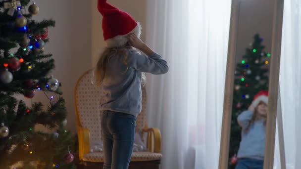 Γοητευτικό λευκό κοριτσάκι που δοκιμάζει χριστουγεννιάτικο καπέλο που ποζάρει μπροστά στον καθρέφτη στο σπίτι. Θετικό παιδί ετοιμάζεται για τον εορτασμό της Πρωτοχρονιάς σε εσωτερικούς χώρους. — Αρχείο Βίντεο