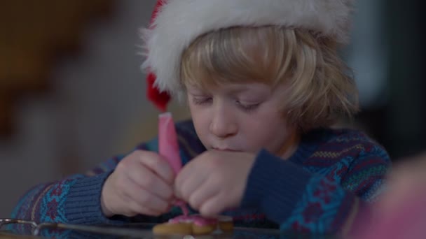 Närbild fokuserad liten pojke dekorera pepparkakor kaka på julafton inomhus hemma. Koncentrerad kaukasiskt barn förbereder läcker söt traditionell dessert på nyår. — Stockvideo