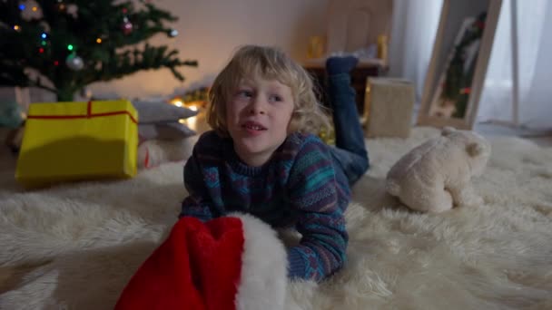 Porträtt av söt vit liten pojke talar leende liggande på mjuk matta i vardagsrummet på julafton. Glad bekymmerslös barn önskar mirakel på Nytt år hemma. Joy och barndomskoncept. — Stockvideo