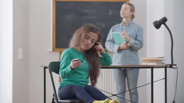 Gelangweilte kleine Person sitzt auf einem Stuhl am Schreibtisch im Klassenzimmer, spielt mit Stift und kreuzt die Hände als verschwommener Lehrer, der im Hintergrund redet. Porträt einer desinteressierten, traurigen Kaukasierin, die drinnen studiert. — Stockvideo