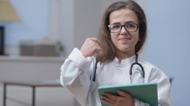 Gözlüklü kendine güvenen zeki kadın doktor portresi başparmağını kaldırıyor ve kameraya bakıyor. Kapalı alanda elinde tabletle. İş yerinde hastanede poz veren mutlu Kafkasyalı küçük kişi.