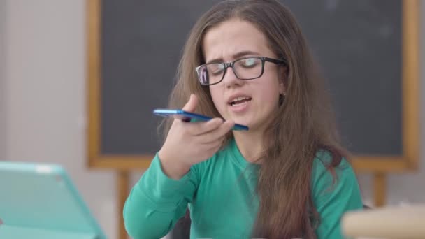 Millennial intelligent liten person talar i smartphone högtalare sitter i universitetets klassrum. Porträtt av smart ung vit kvinna i glasögon inspelning röst meddelande inomhus. — Stockvideo