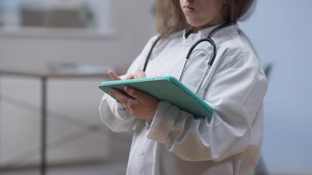 Orang kecil tak dikenal dalam gaun medis berselancar di internet pada tablet yang berdiri di dalam rumah sakit. Wanita kaukasia muda berpikir membaca berita terbaru secara online. Kedokteran dan teknologi modern. — Stok Video