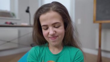 Sağlıklı vitamin elmasını ısıran küçük insan kameranın çiğnediği meyveye gülümsüyor. Kafkasyalı güzel esmer kadının kapalı alandaki sağlıklı yiyeceklerin tadını çıkarırken yakın plan portresi..