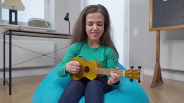 Getalenteerde kleine persoon spelen ukelele zingen zittend op zak stoel binnen thuis. Portret van een zelfverzekerde blanke zangeres die graag lacht. Vrouw rust in het weekend. — Stockvideo
