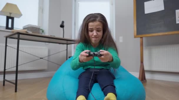 Joueuse se réjouissant de gagner dans un jeu vidéo en ligne assis sur une chaise de sac à l'intérieur. Portrait de petite personne caucasienne excitée utilisant le contrôleur de jeu en regardant la victoire gestuelle de la caméra. — Video