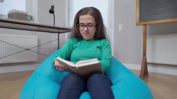 Petite personne intelligente lisant un livre gestuel J'ai eu une idée en souriant assis sur une chaise de sac à l'intérieur. Portrait de génie intelligent confiant femme caucasienne étudiant dans une salle de classe confortable. — Video