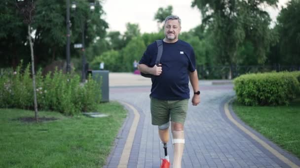 Vista frontal retrato confiante de cabelos grisalhos macho amputado andando em câmera lenta no parque de verão olhando ao redor. Largo tiro homem caucasiano positivo com cabelos grisalhos e pernas protéticas passeando ao ar livre. — Vídeo de Stock