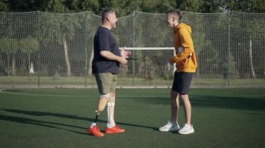 Neşeli, neşeli, olgun bir baba ampüte ve genç bir çocuk açık hava spor sahasında konuşuyor. Neşeli beyaz adam ve çocuk güneşli stadyumda antrenman yapıyor. Yavaş çekim.