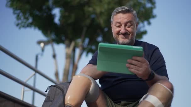 Radostný vousatý muž s amputovaným úsměvem surfující na sociálních sítích na tabletu, který sedí venku. Střední snímek portrét bělocha zralého muže s umělými končetinami a šedivými vlasy těší volný čas. — Stock video