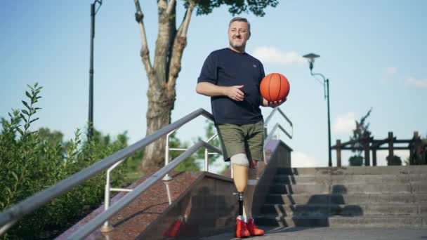 Motivovaný pohledný zralý amputovaný muž s protézou stojící za slunečného dne venku s basketbalovým míčem odvracejícím pohled. Široký portrét bělošského šedovlasého sportovce s umělýma nohama. Zpomalený pohyb — Stock video